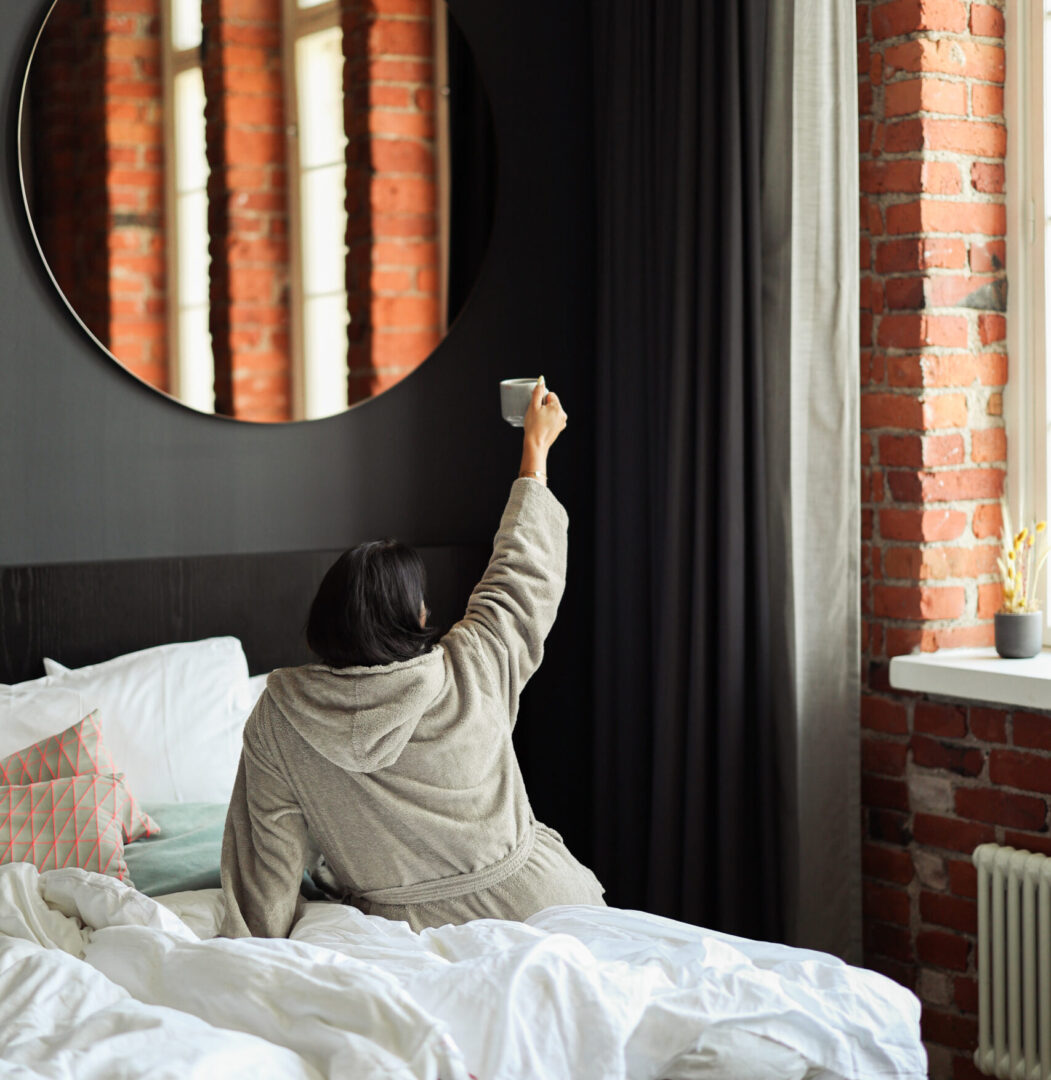 Person in a gray robe sits on a bed, raising a cup toward a round mirror on a dark wall with brick walls and curtains in view behind.