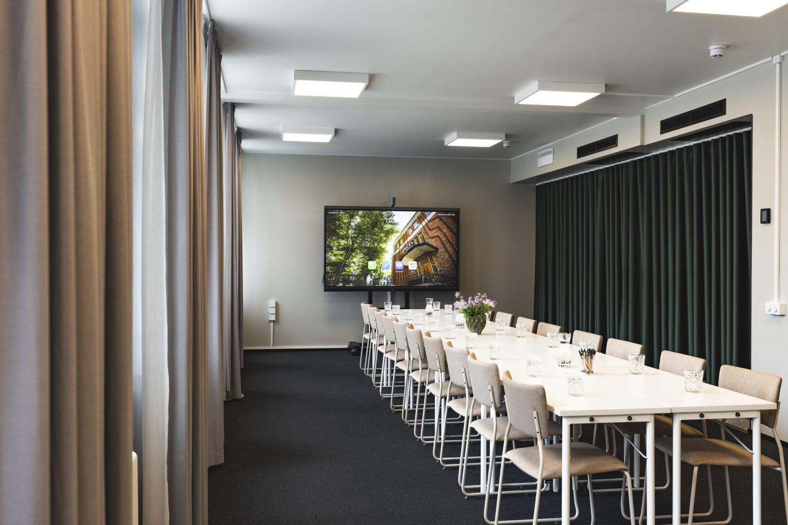 Long conference room table surrounded by beige chairs, with a wall-mounted screen at the far end and thick curtains along the left wall.