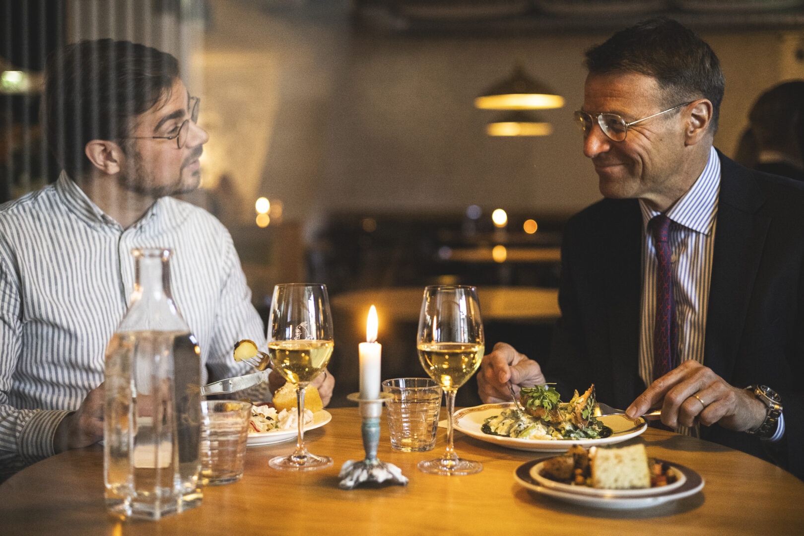 Two men dining at a candlelit restaurant table, glasses of wine in front of them as they talk and smile briefly.