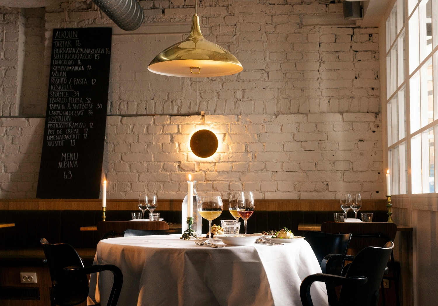 Round table set for dinner with a white tablecloth, candles, and wine glasses in a brick-walled restaurant.