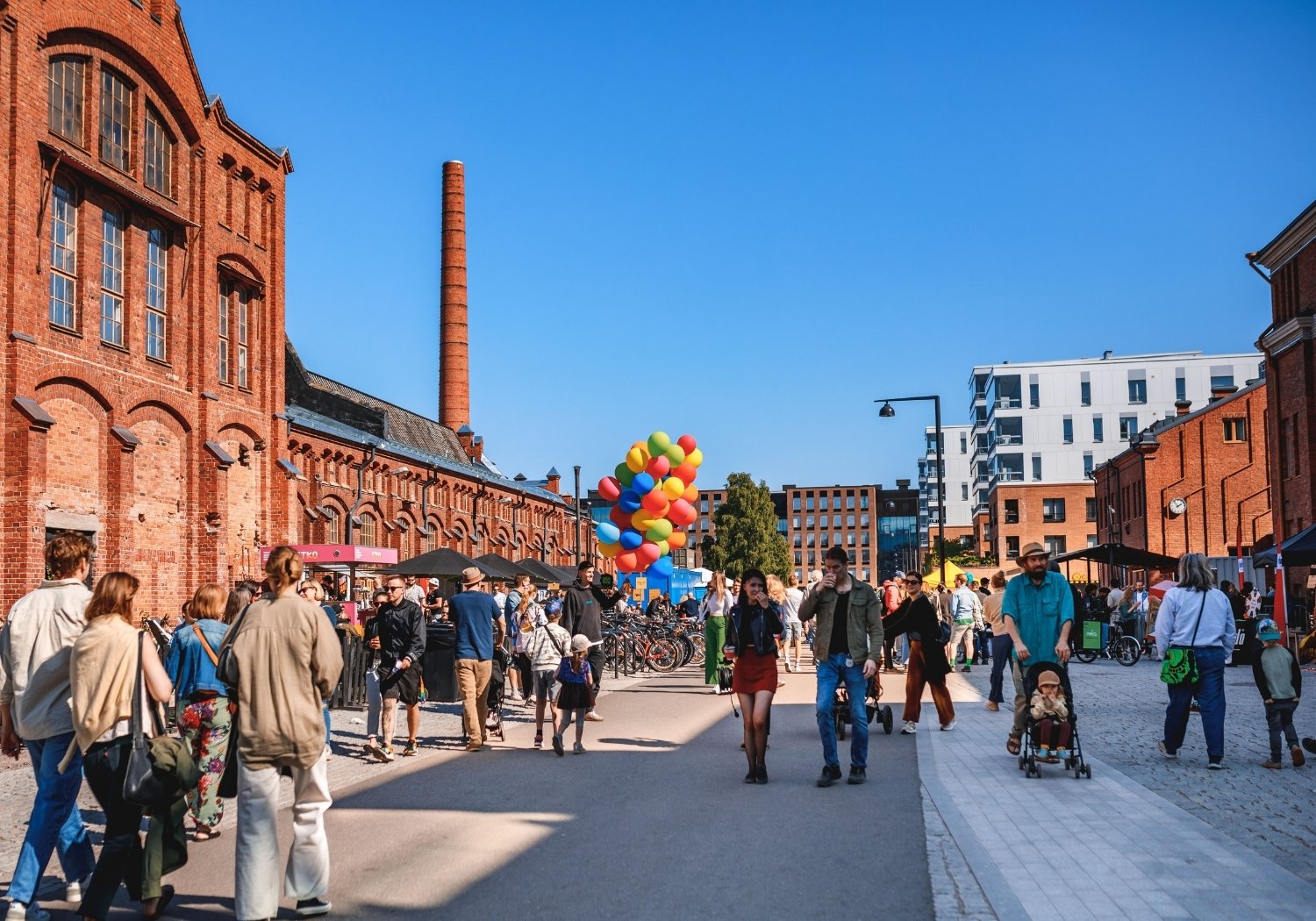 Outdoor street festival in a red brick industrial district with a colorful balloon bouquet centerpiece and crowds strolling along a sunny day.