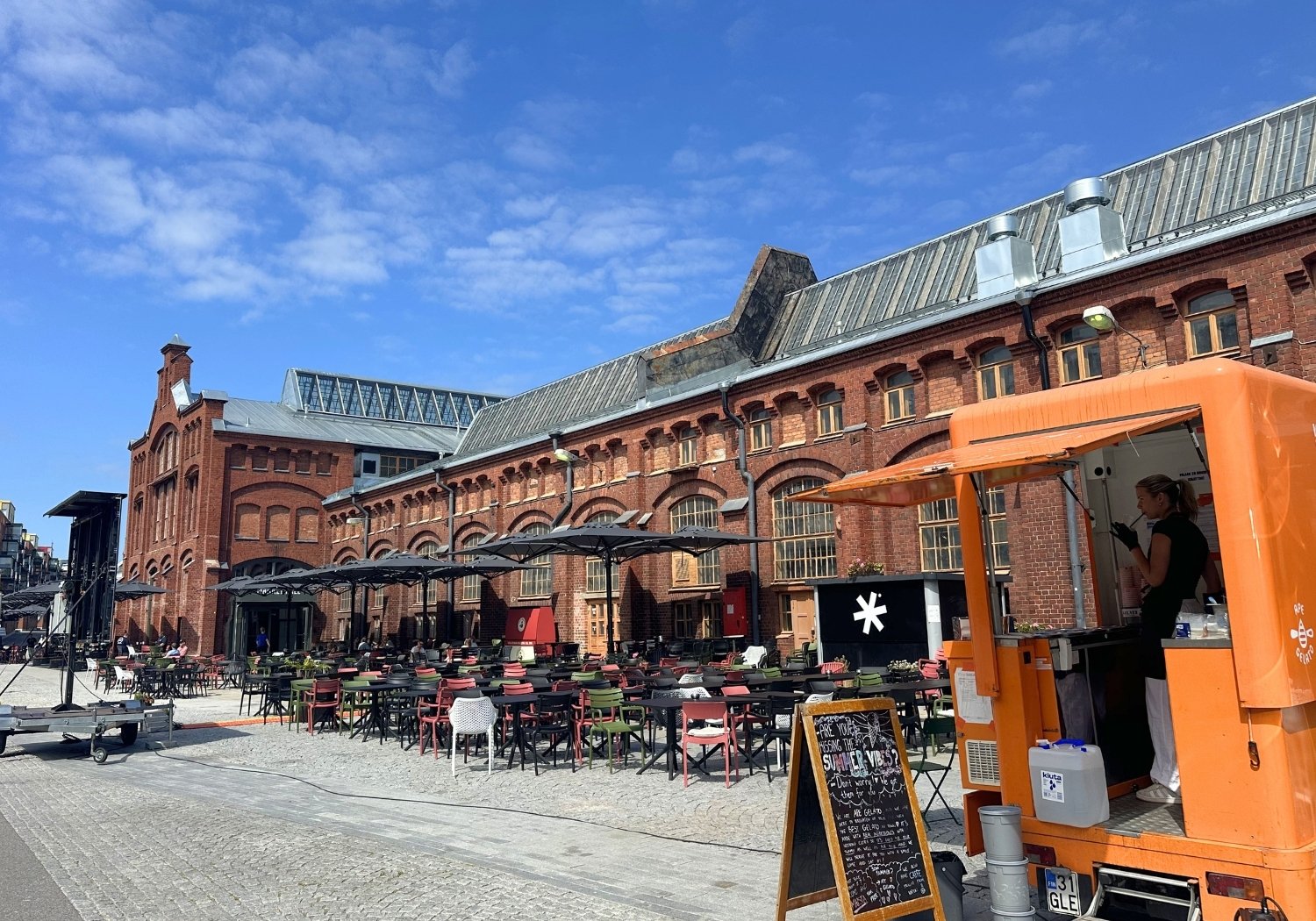 Outdoor cafe setup with multicolored chairs and black umbrellas arranged along a cobblestone square beside a red brick industrial building under a blue sky; an orange food truck with a worker inside stands at right.