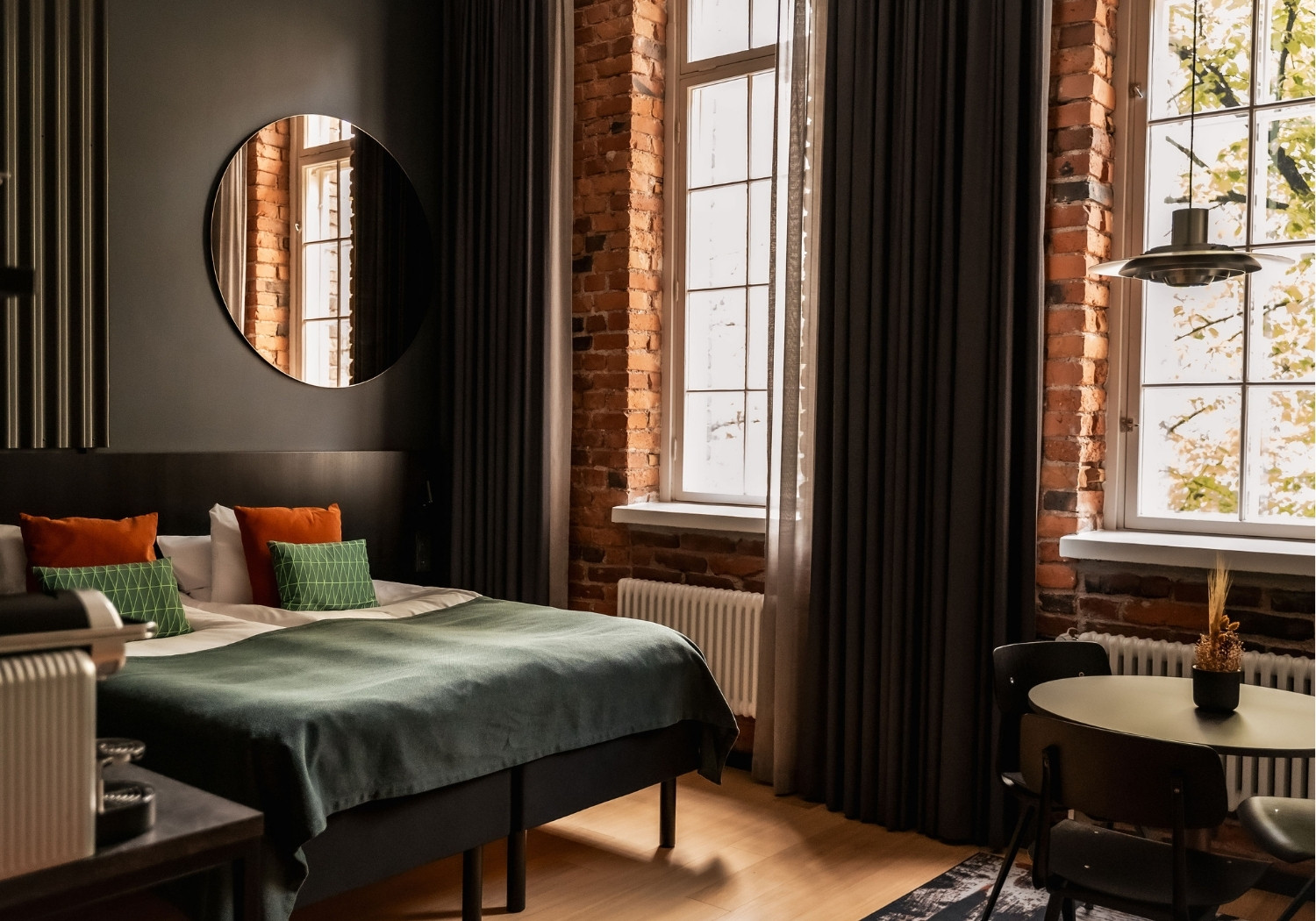 Hotel bedroom with exposed brick walls, a round mirror, dark curtains, and a bed with green and orange cushions.